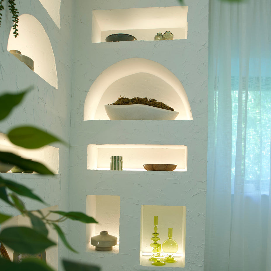 A white stucco wall with built-in arched and rectangular shelves, softly lit from within, holding decorative ceramic vessels and green glassware. Large green leaves and a sheer white curtain frame the scene, with a woven basket visible at the lower right.