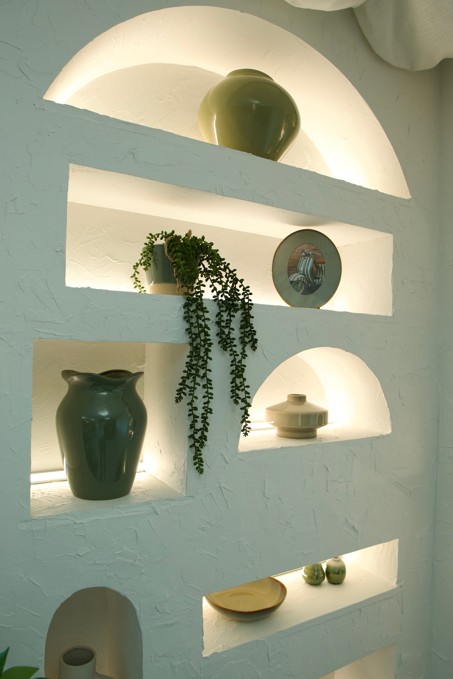 A white textured wall with built-in arched and rectangular shelves illuminated by warm lighting, displaying ceramic vases, a trailing green plant, and decorative plates.